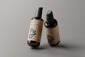 Two amber glass pump bottles are placed against a neutral gray background. The bottles have a minimalistic label featuring the letters PSD and additional text, indicating they are mockup templates. The lighting highlights the glossy finish of the bottles, giving them a professional and sleek appearance.