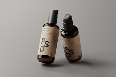 Two amber glass pump bottles are placed against a neutral gray background. The bottles have a minimalistic label featuring the letters PSD and additional text, indicating they are mockup templates. The lighting highlights the glossy finish of the bottles, giving them a professional and sleek appearance.