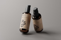 Two amber glass pump bottles are placed against a neutral gray background. The bottles have a minimalistic label featuring the letters PSD and additional text, indicating they are mockup templates. The lighting highlights the glossy finish of the bottles, giving them a professional and sleek appearance.