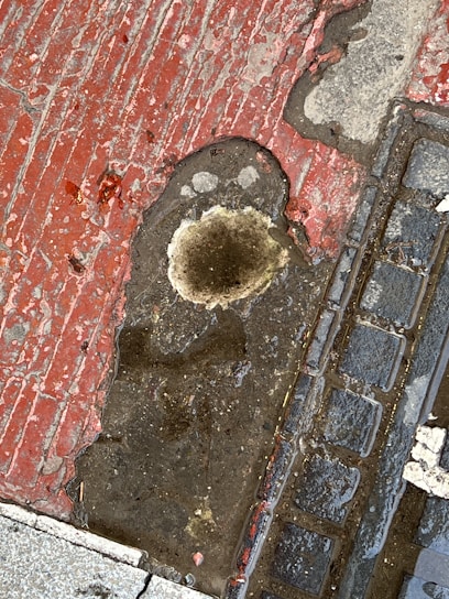 A weathered sidewalk surface shows a mix of red and beige concrete with a prominent irregular hole filled with water. Surrounding it, the surface appears cracked and stained. Adjacent to the hole is a section of cobblestone paving, slightly wet, adding a textured contrast to the scene.