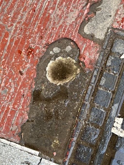 A weathered sidewalk surface shows a mix of red and beige concrete with a prominent irregular hole filled with water. Surrounding it, the surface appears cracked and stained. Adjacent to the hole is a section of cobblestone paving, slightly wet, adding a textured contrast to the scene.