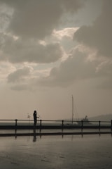 A silhouette of a person stands near a waterfront, holding microphones with a cloudy sky overhead. A sailboat is visible in the background, and there's a soft reflection on the wet ground.