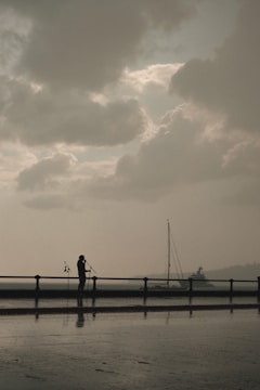 A silhouette of a person stands near a waterfront, holding microphones with a cloudy sky overhead. A sailboat is visible in the background, and there's a soft reflection on the wet ground.