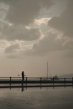 A silhouette of a person stands near a waterfront, holding microphones with a cloudy sky overhead. A sailboat is visible in the background, and there's a soft reflection on the wet ground.