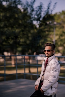 A person wearing sunglasses and a branded white puffer jacket leans casually against a metal railing. The background includes blurred greenery and a sunny outdoor setting.