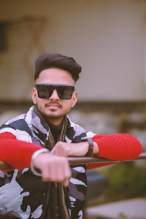A person wearing large sunglasses and a stylish outfit, including a camo-patterned jacket and a red sweater, appears relaxed while resting their arm on a railing. The background is blurred, suggesting an outdoor setting.