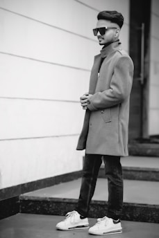A person stands confidently outdoors, wearing a stylish coat, oversized sunglasses, and sneakers. They have a trendy hairstyle and are positioned in front of a modern building facade.