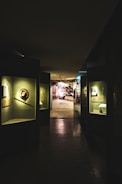 A dimly lit museum hallway with various exhibits displayed in glass cases along the walls. The exhibits are illuminated by individual lights, casting shadows on the polished floor. At the end of the hallway, there is a brightly lit area with a photograph or mural of an industrial or historical scene.