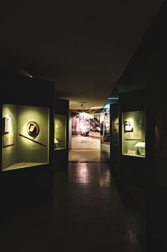 A dimly lit museum hallway with various exhibits displayed in glass cases along the walls. The exhibits are illuminated by individual lights, casting shadows on the polished floor. At the end of the hallway, there is a brightly lit area with a photograph or mural of an industrial or historical scene.