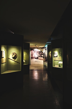 A dimly lit museum hallway with various exhibits displayed in glass cases along the walls. The exhibits are illuminated by individual lights, casting shadows on the polished floor. At the end of the hallway, there is a brightly lit area with a photograph or mural of an industrial or historical scene.