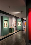 A gallery hallway with artworks displayed on the walls. There are green display frames featuring various sculptures or reliefs, and an enclosed glass display with an artifact. The floor is polished concrete, and the lighting is soft, highlighting the artwork.