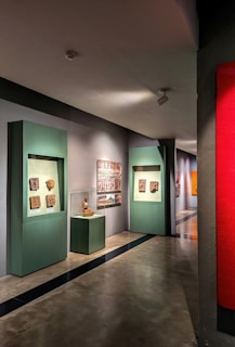 A gallery hallway with artworks displayed on the walls. There are green display frames featuring various sculptures or reliefs, and an enclosed glass display with an artifact. The floor is polished concrete, and the lighting is soft, highlighting the artwork.