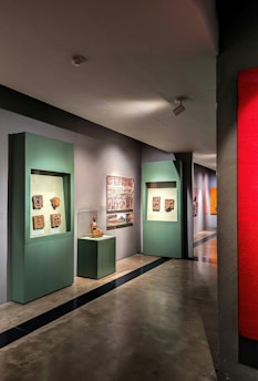 A gallery hallway with artworks displayed on the walls. There are green display frames featuring various sculptures or reliefs, and an enclosed glass display with an artifact. The floor is polished concrete, and the lighting is soft, highlighting the artwork.