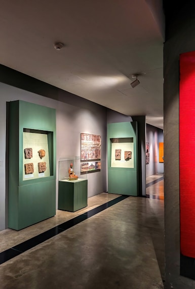 A gallery hallway with artworks displayed on the walls. There are green display frames featuring various sculptures or reliefs, and an enclosed glass display with an artifact. The floor is polished concrete, and the lighting is soft, highlighting the artwork.