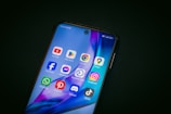 A smartphone displays a variety of popular app icons on its screen, including YouTube, Google Play Store, Chrome, Google Maps, Facebook, Messenger, Business Suite, Instagram, WhatsApp, Pinterest, Discord, and TikTok. The screen has a vibrant background with blue and purple hues.