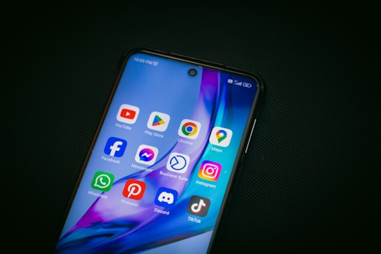 A smartphone displays a variety of popular app icons on its screen, including YouTube, Google Play Store, Chrome, Google Maps, Facebook, Messenger, Business Suite, Instagram, WhatsApp, Pinterest, Discord, and TikTok. The screen has a vibrant background with blue and purple hues.