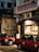 Two red taxis are parked on a dimly lit street beside a building with signage in Chinese characters. A person wearing a white jacket is approaching the entrance of a shop or restaurant. The building's exterior is slightly worn, with visible air conditioning units.