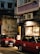 Two red taxis are parked on a dimly lit street beside a building with signage in Chinese characters. A person wearing a white jacket is approaching the entrance of a shop or restaurant. The building's exterior is slightly worn, with visible air conditioning units.