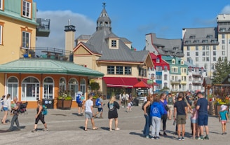A bustling street scene in a vibrant, European-style village with colorful buildings. People, including families and groups, are walking and socializing in the open plaza.