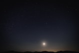 A night sky filled with stars above a silhouetted mountain range. A bright celestial body, possibly the moon or a planet, illuminates the scene from near the horizon.