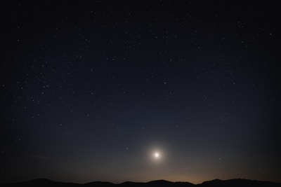 A night sky filled with stars above a silhouetted mountain range. A bright celestial body, possibly the moon or a planet, illuminates the scene from near the horizon.
