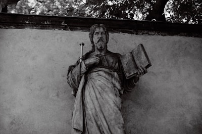 A stone statue of a bearded figure holding a book in one hand and a staff in the other. The statue is set against a textured wall with foliage visible above.