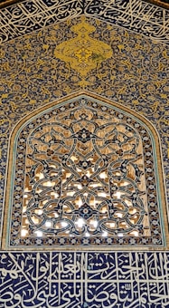 Intricate Islamic geometric patterns are displayed on a wall with elaborate blue and gold tilework. The design features ornate floral and geometric motifs, along with Arabic calligraphy. The craftsmanship showcases meticulous detail and artistry.