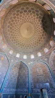An intricately designed mosque dome featuring detailed geometric and floral patterns with alternating colored tiles. The blue and gold hues create a mesmerizing effect, emphasizing the architectural symmetry and cultural richness of the structure.
