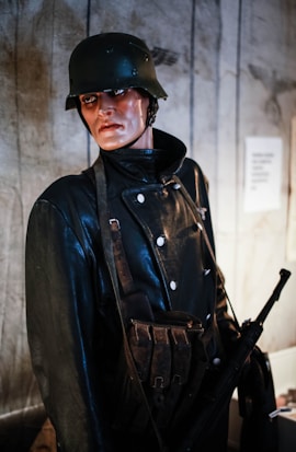 A mannequin dressed in a soldier's uniform, featuring a dark leather coat with double-breasted buttons and a matching helmet. The figure is equipped with a shoulder strap and ammunition pouches, while holding a rifle. The background includes wooden panels and a partially visible sign.