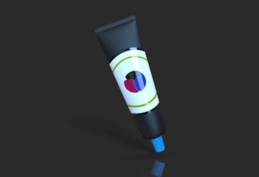 A black tube with a cylindrical shape is standing upright against a dark background. It features a white label with colorful abstract shapes including red, blue, and black. The cap of the tube is blue.