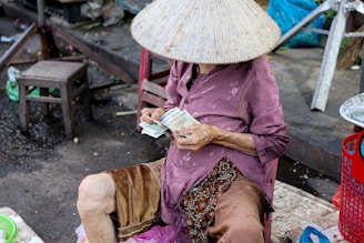 A person wearing a traditional conical hat is seated on a stool while counting paper currency. The surroundings suggest a market setting with a few chairs scattered around. The individual is dressed in a purple shirt and brown shorts, and appears to be older given the visible hand wrinkles.