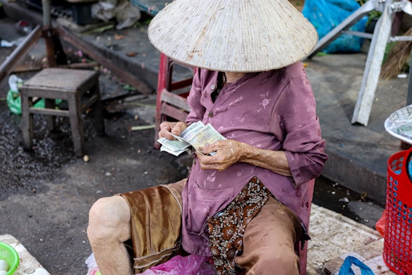 A person wearing a traditional conical hat is seated on a stool while counting paper currency. The surroundings suggest a market setting with a few chairs scattered around. The individual is dressed in a purple shirt and brown shorts, and appears to be older given the visible hand wrinkles.
