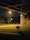 A shopping cart is positioned under a bridge at night, with a streetlamp casting a warm glow on the nearby path and grass. The environment includes a railway track parallel to the path and large stone structures, with trees in the distance.