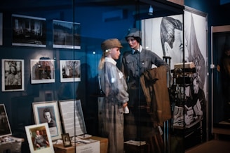 The image features a museum exhibit displaying mannequins dressed in historical military uniforms. Various framed photographs and documents are showcased on the walls and in display cases. A large poster or mural of a historical figure is visible in the background.