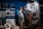 The image features a museum exhibit displaying mannequins dressed in historical military uniforms. Various framed photographs and documents are showcased on the walls and in display cases. A large poster or mural of a historical figure is visible in the background.