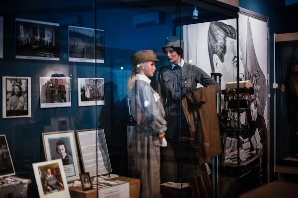 The image features a museum exhibit displaying mannequins dressed in historical military uniforms. Various framed photographs and documents are showcased on the walls and in display cases. A large poster or mural of a historical figure is visible in the background.