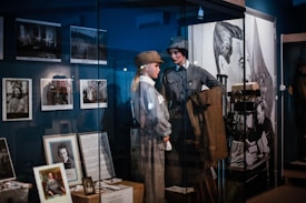 The image features a museum exhibit displaying mannequins dressed in historical military uniforms. Various framed photographs and documents are showcased on the walls and in display cases. A large poster or mural of a historical figure is visible in the background.