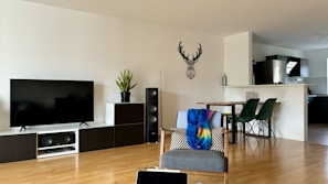 A modern living room with a large flat-screen TV on a black and white TV stand, accompanied by a potted plant and speakers. A gray chair draped with a colorful blanket and cushion is in the foreground. A dining area with a wooden table and green chairs is visible behind the chair. The kitchen is partially visible, separated by a small partition. A decorative wall piece in the shape of a deer's head adorns the wall.