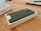 A sleek, dark-colored smartphone lies within an open white box on a wooden table. The phone's camera lenses are prominent, and there is a rectangle of white paper or plastic over part of the phone. Nearby, a notebook and pen are partially visible.
