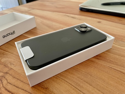 A sleek, dark-colored smartphone lies within an open white box on a wooden table. The phone's camera lenses are prominent, and there is a rectangle of white paper or plastic over part of the phone. Nearby, a notebook and pen are partially visible.