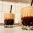 Two glass tumblers filled with a layered beverage, featuring a dark bottom and a frothy, lighter top. Each glass has a black straw for sipping and intricate faceted designs on the surface. The glasses rest on a light-colored countertop in front of a white tiled wall, with some small kitchen utensils like toothpicks in the background.