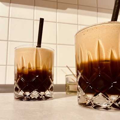 Two glass tumblers filled with a layered beverage, featuring a dark bottom and a frothy, lighter top. Each glass has a black straw for sipping and intricate faceted designs on the surface. The glasses rest on a light-colored countertop in front of a white tiled wall, with some small kitchen utensils like toothpicks in the background.