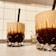 Two glass tumblers filled with a layered beverage, featuring a dark bottom and a frothy, lighter top. Each glass has a black straw for sipping and intricate faceted designs on the surface. The glasses rest on a light-colored countertop in front of a white tiled wall, with some small kitchen utensils like toothpicks in the background.
