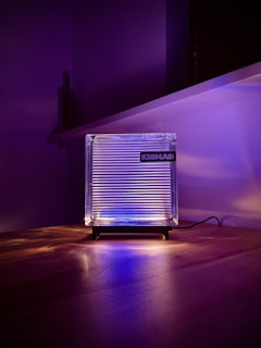A square, clear acrylic or glass lamp is placed on a wooden surface with a black stand. It emits a soft glow of purple and blue light, casting subtle reflections and shadows on the table. The word 'KISMAS' is visible on the lamp. The background is a dimly lit room with purple lighting, creating a modern and minimalistic atmosphere.