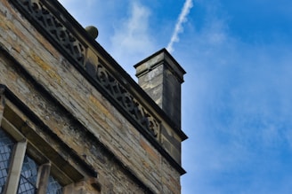 A close-up view of an old stone building's exterior, featuring a decorative stone railing with intricate patterns. The building is adorned with a tall chimney rising against a vivid blue sky with wispy clouds.
