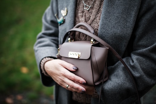 A person is wearing a gray coat and holding a small, elegant brown handbag with a gold clasp. They have neatly manicured nails, painted in a neutral shade, and are wearing a knitted sweater underneath the coat. There are decorative pins on the coat, including a butterfly and a constellation. The background is a blur of green foliage, suggesting a natural outdoor setting.