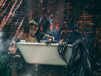 A creature with a sinister appearance and scaly skin is emerging from an old-fashioned bathtub in a strange, eerie setting. The creature has a crown of leaves and flowers on its head and is reaching out with clawed hands. The backdrop includes a brick wall partially obscured by plants and flowers, with starfish and other decorations hanging. There's an aura of mist and smoke, enhancing the mysterious atmosphere.
