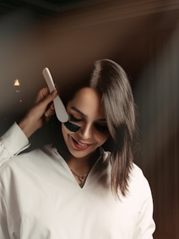 A person with short, dark hair is smiling while a hand applies a black facial mask with a spatula to their cheek. They are wearing a white top and a necklace. The background is softly blurred, creating a warm and relaxed atmosphere.