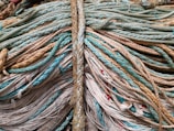 An array of ropes of varying thicknesses and colors intertwined and laid out in an overlapping fashion. The ropes range in color from teal and beige to off-white and light brown, with some sections showing red protrusions. The texture is prominent, highlighting the woven nature of the ropes.