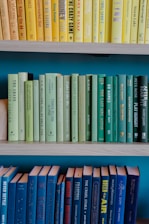 A series of books arranged on three shelves. The top shelf contains books with yellow covers, the middle shelf features books with green covers, and the bottom shelf holds books with blue covers. The shelves create a gradient effect with the color of the book spines.